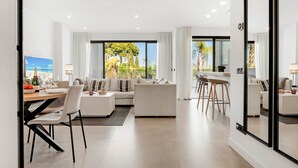 A modern living room with a sofa, dining area, and a large window with a view of palm trees. - Luxury Mirador Resort Apartment (Estepona)
