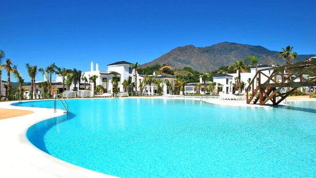 A large, clear blue pool with a curved edge, surrounded by white buildings and palm trees, with a mountain in the background. - Luxury Mirador Resort Apartment (Estepona)