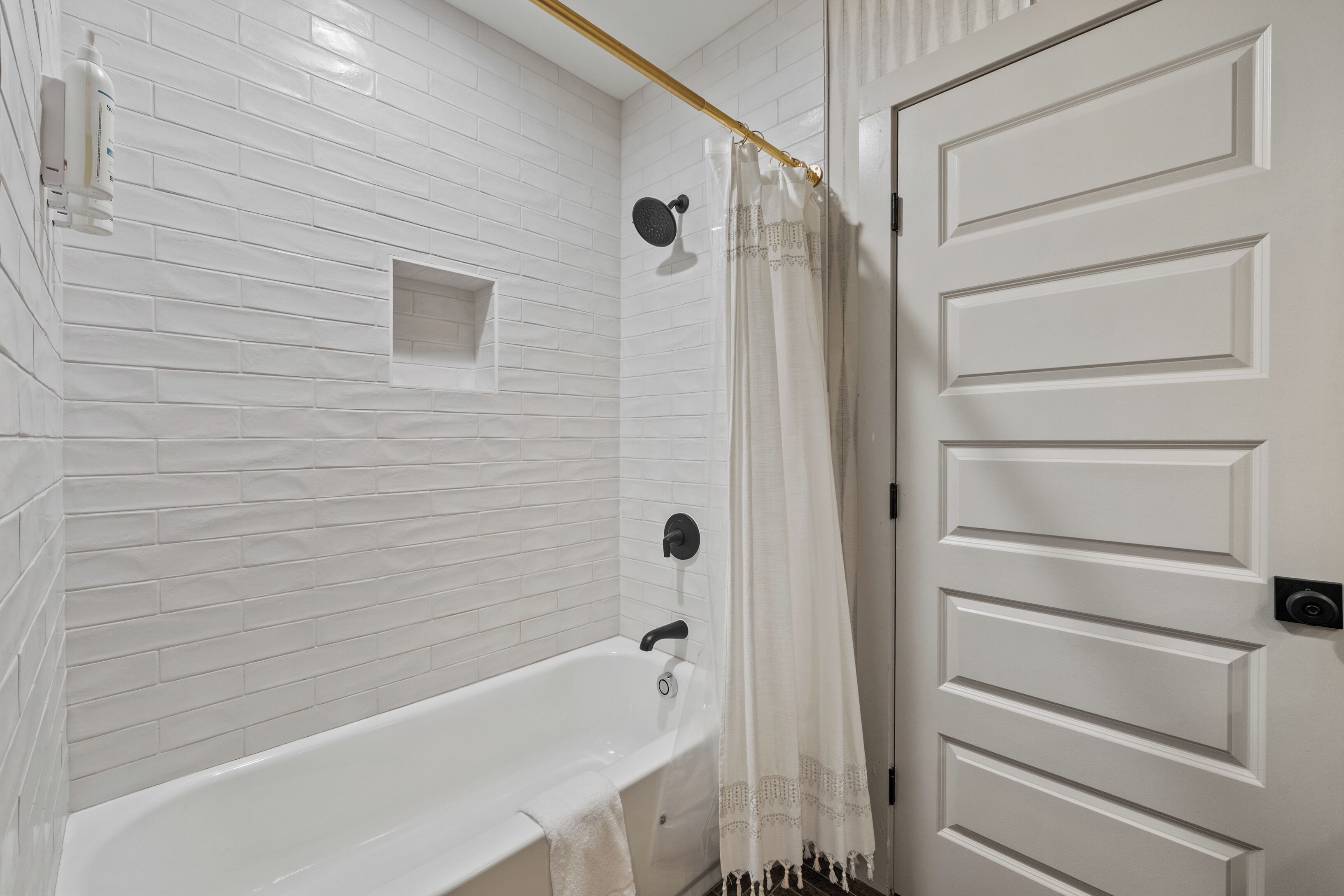 A bathroom with white tiled walls, a white bathtub, a shower with a black handheld showerhead, and a white door with a gold handle.