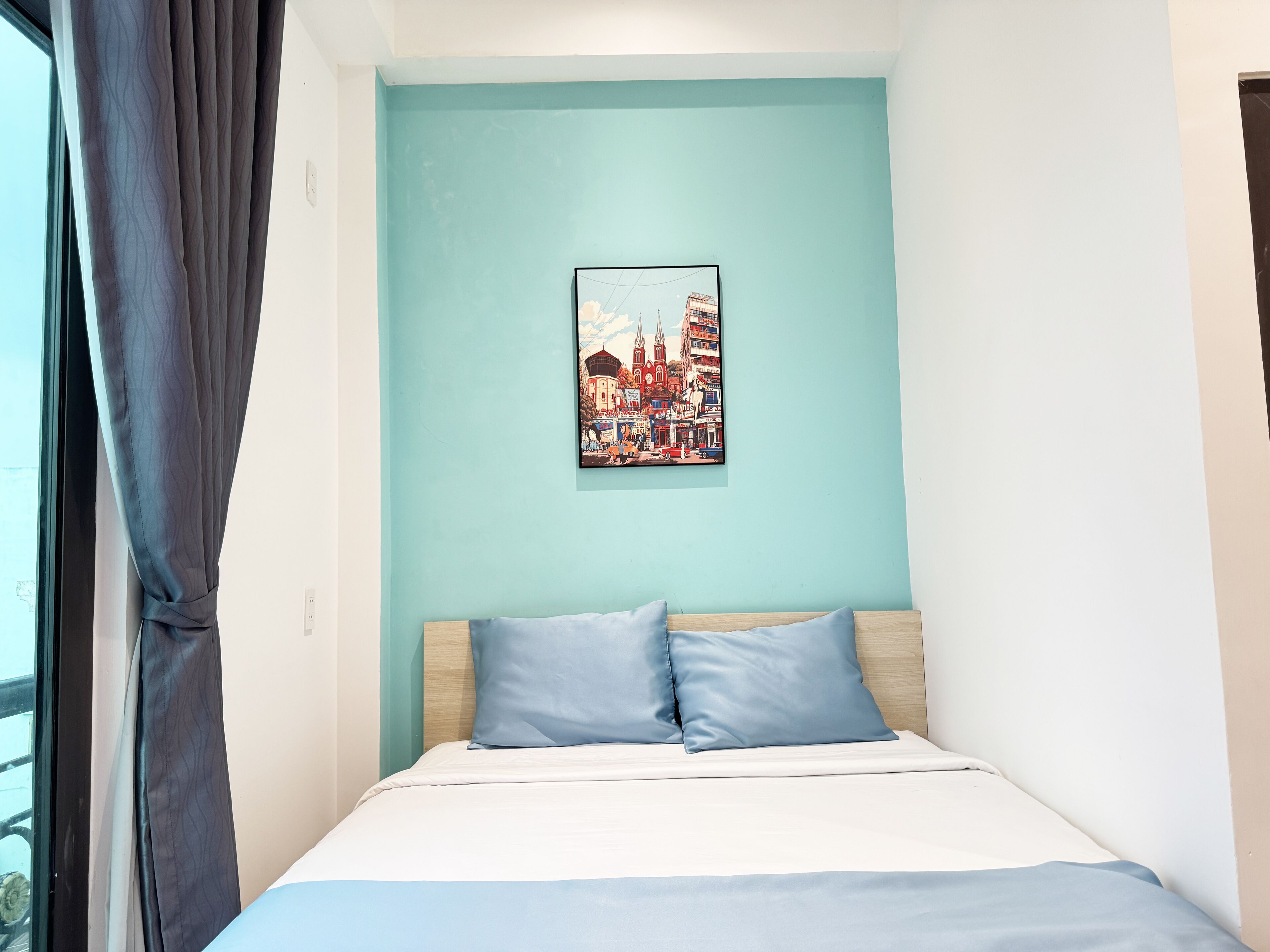 A bedroom with a bed, two pillows, a light blue wall, and a cityscape painting.