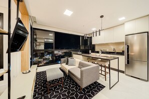 A modern kitchen with a large refrigerator, a dining area with a sofa and a table, and a view of the city at night. - Studio H Residencial Flipchave STH000 (Joinville)