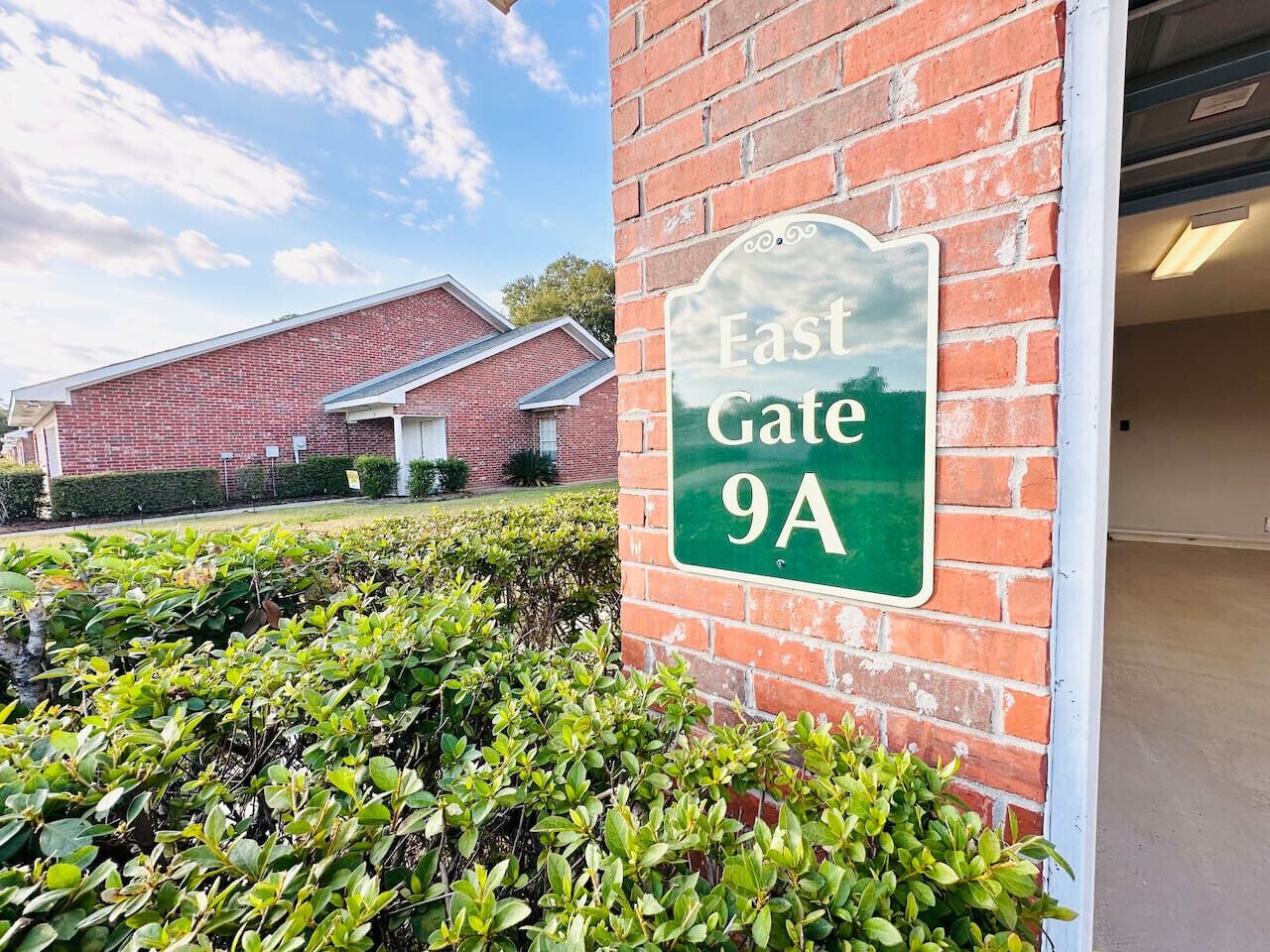 A sign for 'East Gate 9A' on a brick wall, with a building in the background and well-maintained bushes in the foreground.