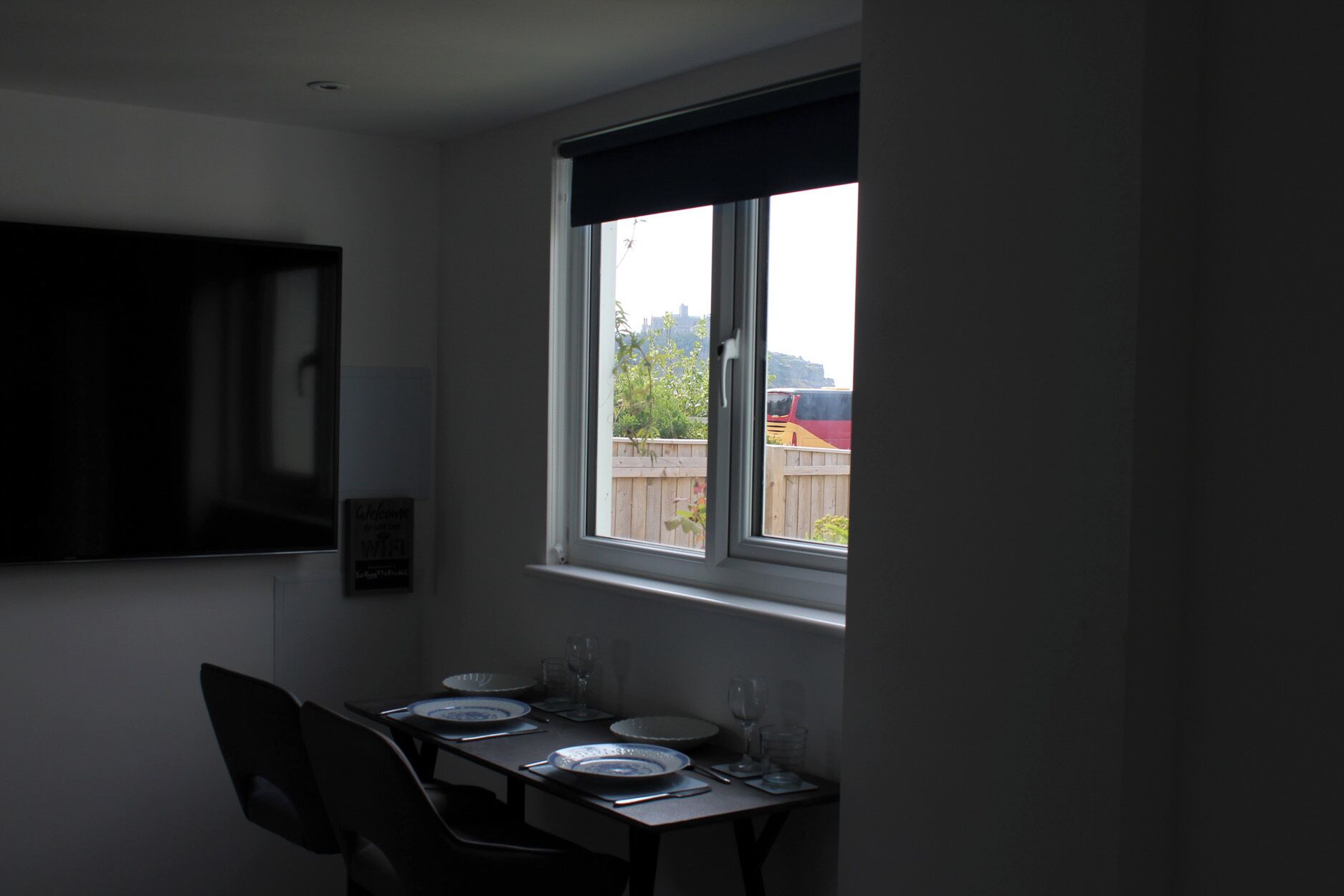 Un comedor con una mesa dispuesta para dos personas, una ventana con vista a un vehículo afuera y una lámpara fijada a la pared.