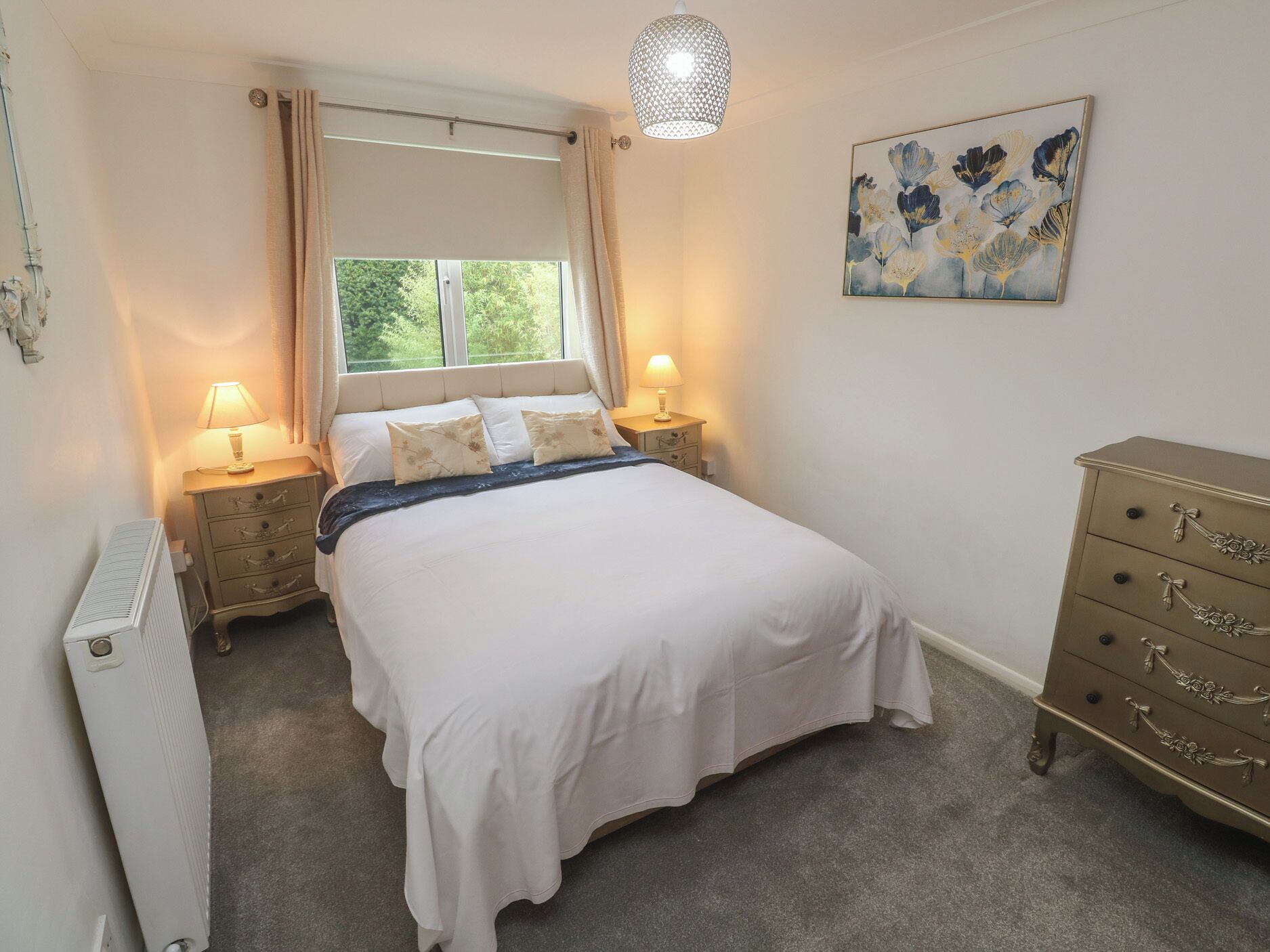 Une chambre à coucher avec un lit, deux lampes de chevet, un radiateur, une commode et une fenêtre avec des rideaux.