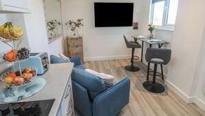 Uma cozinha compacta com um sofá azul, uma área de jantar com duas cadeiras, uma mesa pequena e uma TV fixada na parede.