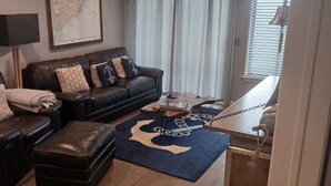 A living room with a dark leather sofa, matching ottoman, and a blue rug with a white anchor design. A wooden coffee table with a book and a decorative bell hangs on the wall. - 2-bedroom condo in wonderful Isle of Palms with AC
Beach and Pier access (Isle of Palms)