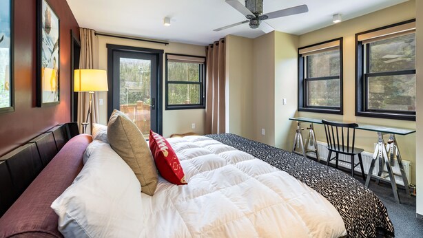 A hotel room with a bed, a desk, a chair, a ceiling fan, and large windows. - Bavarian Chalet - Ski-In Ski-Out Taos Ski Valley (Taos Ski Valley)
