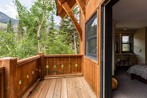 A wooden deck with a view of a forest and mountains, a bedroom with a bed and desk, and a large window.