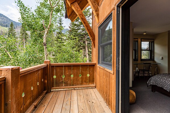 A wooden deck with a view of a forest and mountains, a bedroom with a bed and desk, and a large window.