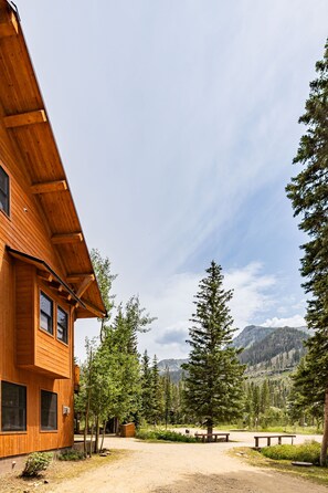 A wooden cabin with a dirt ground, surrounded by trees and mountains in the background.