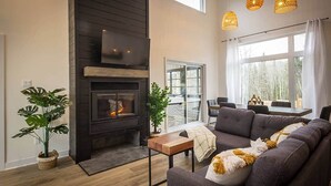 A modern living room with a fireplace, a sofa, a coffee table, and a dining area with a view. - Le Cozy – Hot Tub, Beach & Ski Getaway Tremblant (Lac-Supérieur)