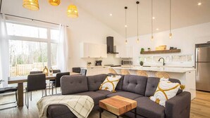 A modern living room with a grey sectional sofa, a wooden coffee table, and a dining area with chairs. There is a kitchen with white cabinets and a stainless steel refrigerator. The room has large windows and hanging pendant lights. - Le Cozy – Hot Tub, Beach & Ski Getaway Tremblant (Lac-Supérieur)