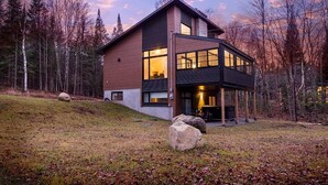 A modern two-story house with large windows and a covered porch, surrounded by a forested area. - Le Cozy – Hot Tub, Beach & Ski Getaway Tremblant (Lac-Supérieur)