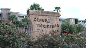 Un segnale con il testo 'Grand Caribbean Coast' circondato da vegetazione lussureggiante e cespugli fioriti.