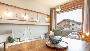 Ett modernt vardagsrum med ett matbord i trä, en soffa och ett stort fönster med utsikt över en byggnad.