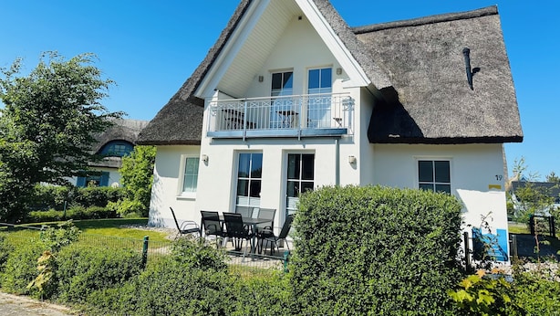 Une maison à deux étages dotée d’un balcon, d’un toit de chaume et d’un jardin bien entretenu.