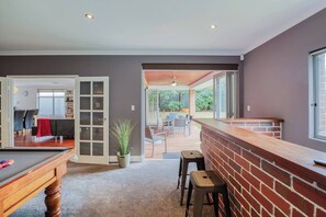 Um quarto amplo com mesa de bilhar, área de bar com banquetas e vista para um pátio externo com móveis.