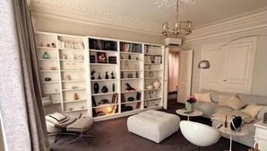 Ein geräumiger Raum mit einer großen, weißen Bücherwand, einer gemütlichen Sitzecke inklusive Sofa und Hocker sowie einer dekorativen Deckenleuchte.
