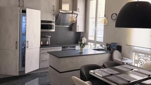 Eine moderne Küche mit einer zentralen Kochinsel, dunklen Arbeitsplatten und hellen Küchenschränken. Es gibt einen Essbereich mit Tisch und Stühlen sowie Pendelleuchten darüber.