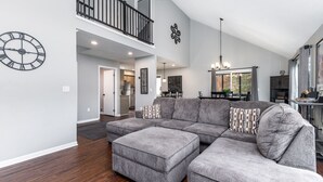A spacious living room with a sectional sofa, a large wall clock, and a balcony with a railing. - 3BdRoom Home w/ Hot Tub + Game Room (Galena)