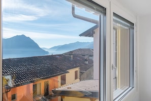 Blick durch ein Fenster auf ein Gebäude mit Ziegeldach, einem Balkon und einer Berglandschaft im Hintergrund.