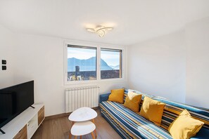 Ein modernes Wohnzimmer mit einem großen Flachbildfernseher, einem Sofa mit gelben Kissen, einem weißen Tisch und einem Fenster mit Blick auf die Berge.