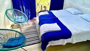 A bedroom with a bed, two blue curtains, a window, a chair, and a rug. - Zeta Camping (Juta)