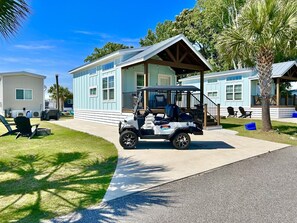 A golf cart parked in front of a building with a porch, a lawn with chairs, and a palm tree. - Cozy Coastal Cottage #61 (North Myrtle Beach)