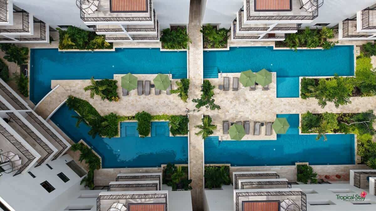 A rooftop pool area with multiple lounge chairs and umbrellas, surrounded by greenery and modern architecture.