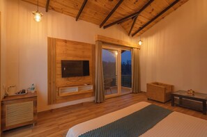 Une chambre avec un plafond en bois, un téléviseur à écran plat fixé sur un panneau en bois, une porte-fenêtre coulissante donnant sur un balcon et une petite table avec une lampe.