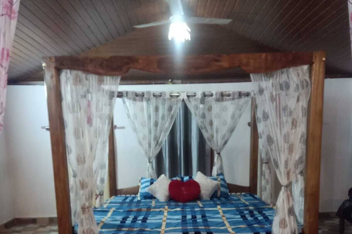 Un lit à baldaquin avec une couvre-lit à carreaux, un oreiller rouge et une tête de lit en bois. Un ventilateur de plafond est installé au-dessus, et des rideaux à motifs ornent les fenêtres.
