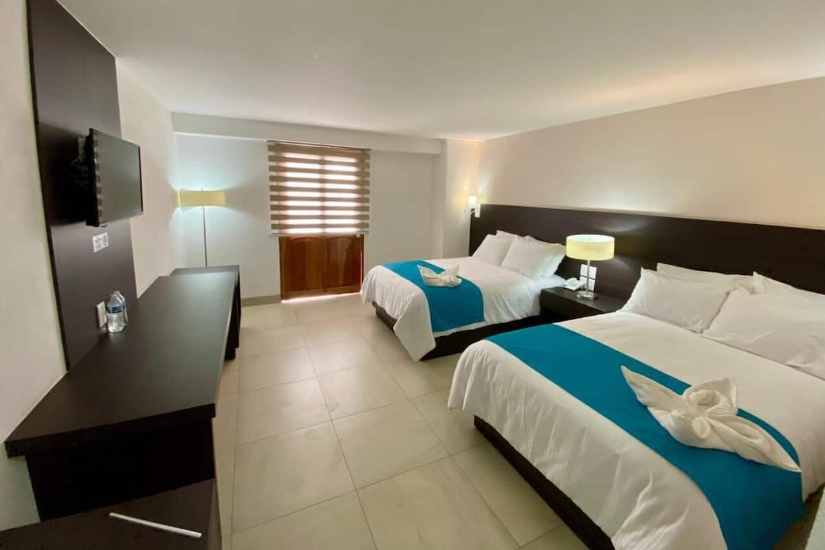 A hotel room with two beds, a desk, a TV, and a window with blinds.