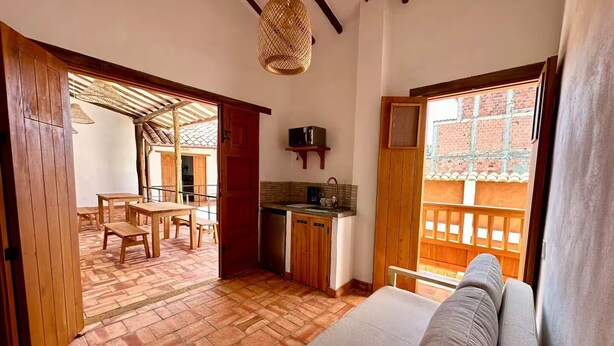 A cozy room with a wooden floor, a sofa, a dining table with chairs, a kitchen area, and a view of a brick building through a window. - Casa Monica - Apt Astromelia (Barichara)