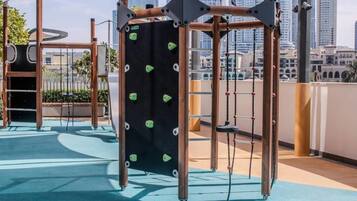 A rooftop playground with climbing structures and swings, surrounded by a cityscape.
