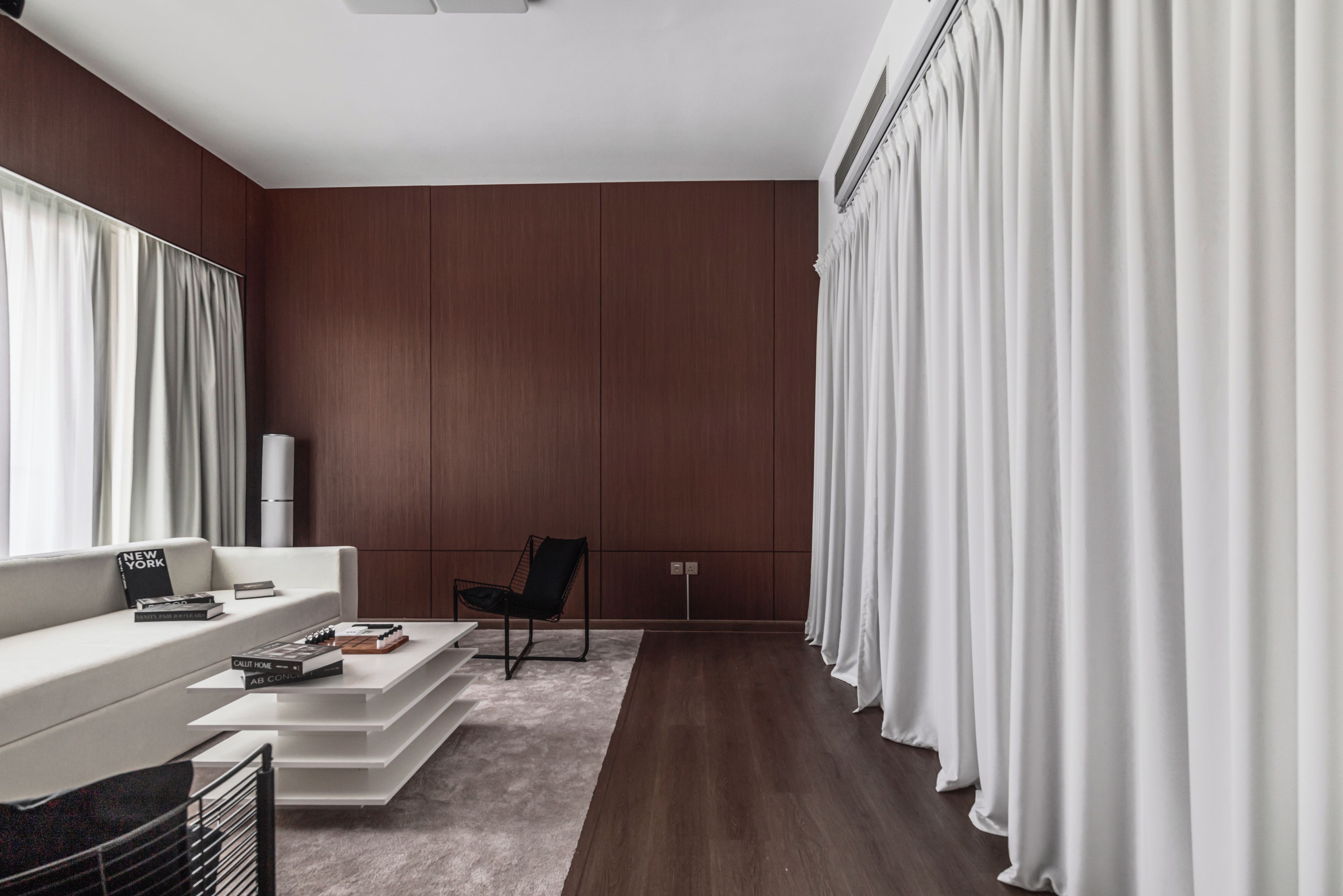 Un salon moderne avec un canapé blanc, un fauteuil noir et une table basse. Il y a une grande fenêtre avec des rideaux blancs, une lampe blanche et un plancher en bois.