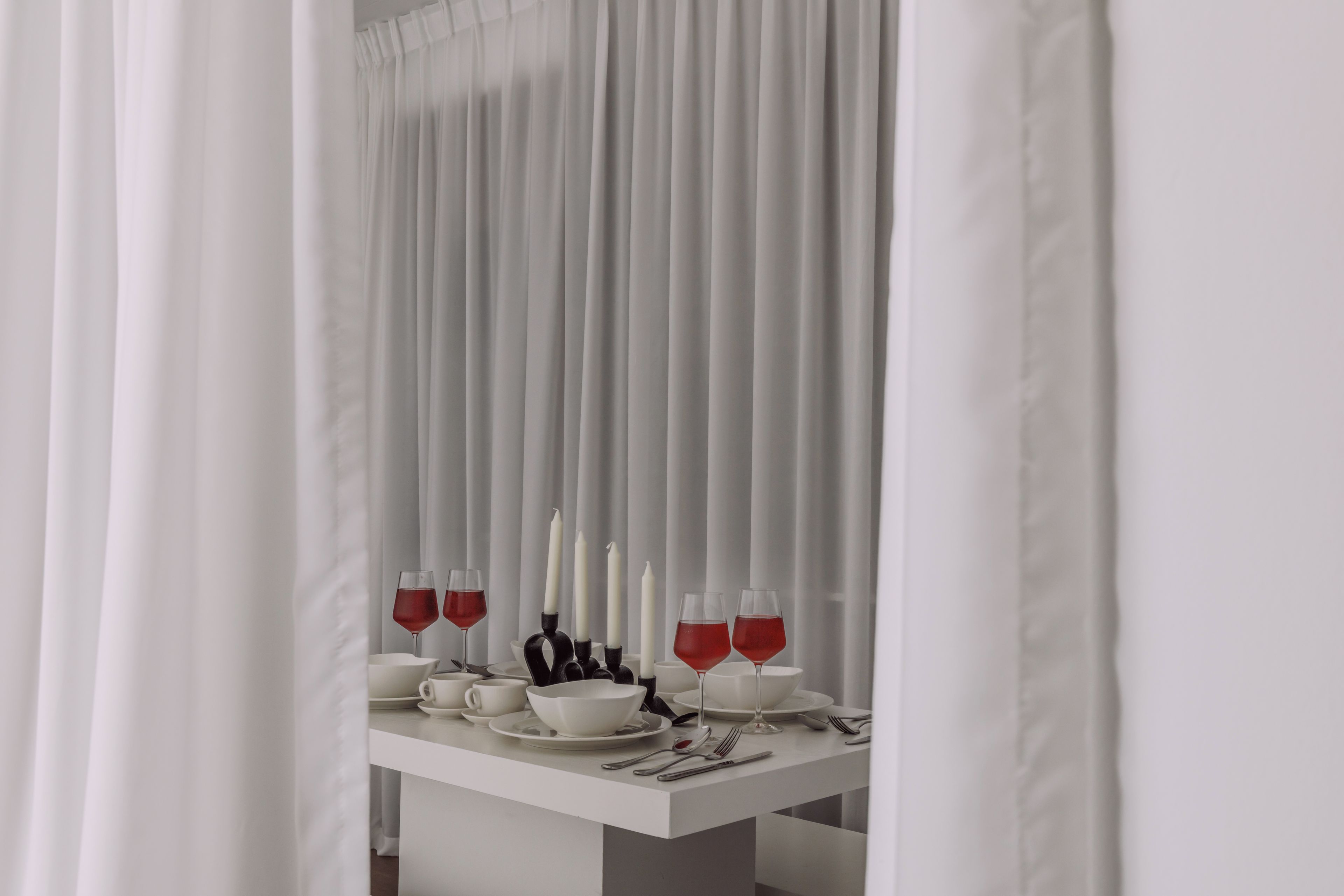 A dining table set with white tableware and red wine glasses, with candles and a white curtain in the background.