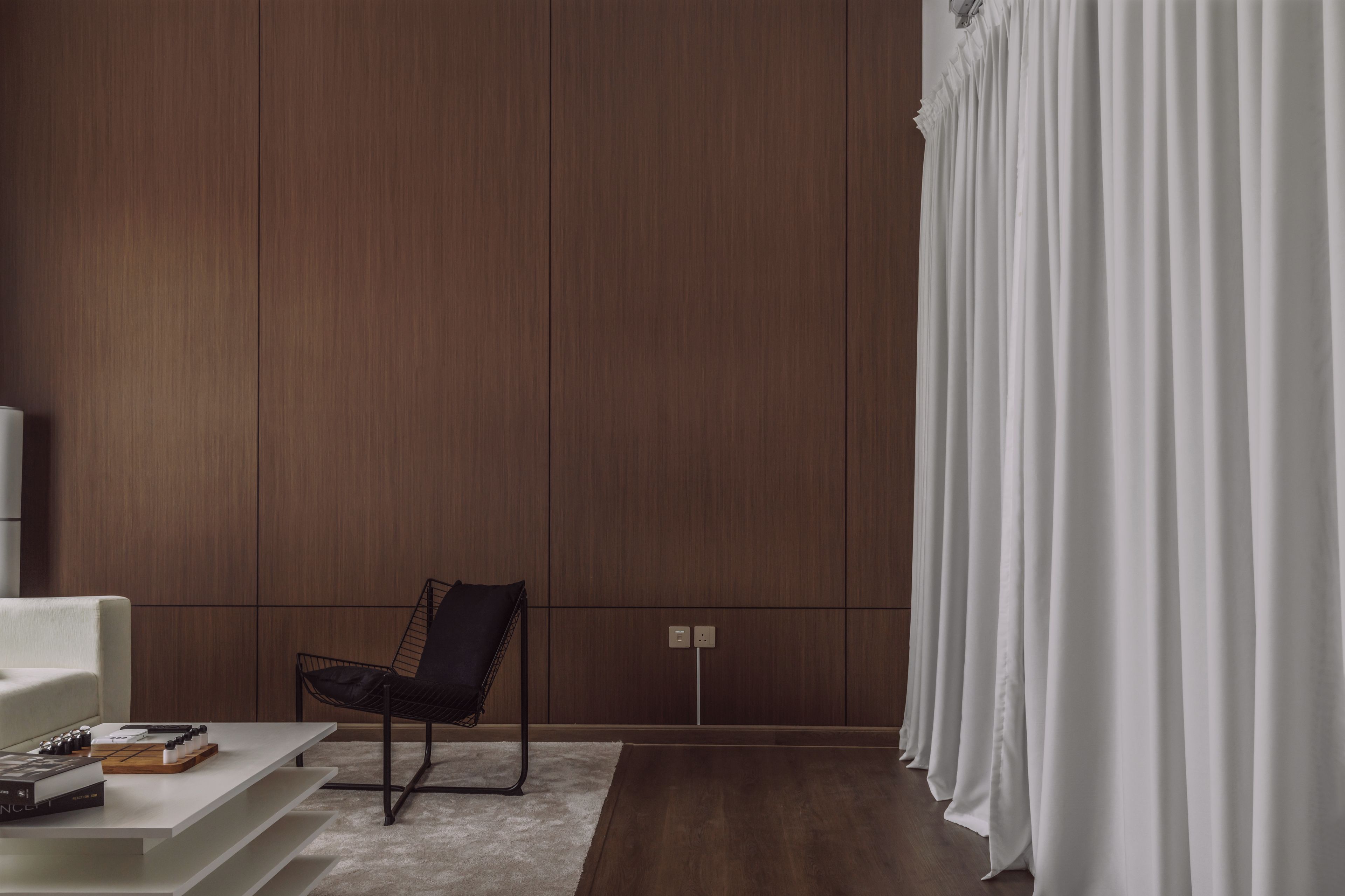 A modern living room with a dark wood panel wall, a black chair, a white sofa, and a white curtain.