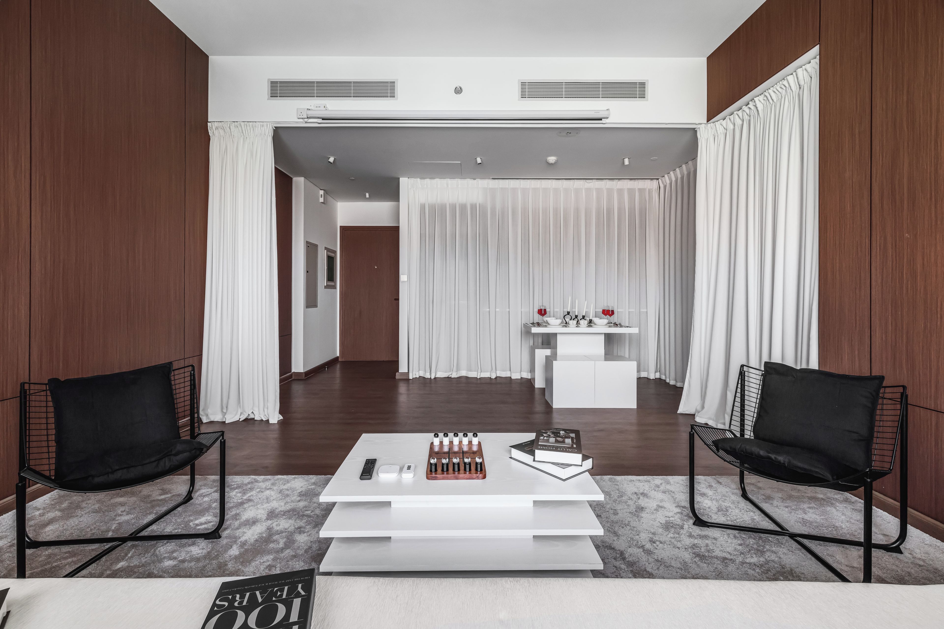 Een moderne woonkamer met een witte salontafel, twee zwarte stoelen, een witte console met decoratieve objecten en een doorkijk naar een andere kamer via een deuropening.