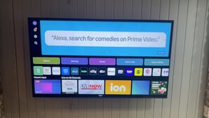 A flat-screen TV mounted on a wall with a grid of colorful app icons, including a search bar with the text "Alexa, search for comedies on Prime Video."