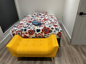 Quarto com uma cama coberta por um edredom estampado com motivos florais, uma poltrona amarela e uma janela com persianas.