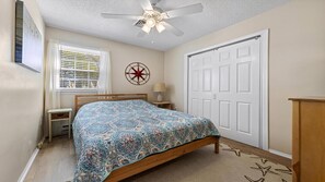 Ein Schlafzimmer mit Bett, Deckenventilator, Fenster mit Jalousien, Tür und Wandbild.