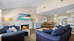 Ein Wohnzimmer mit Kamin, blauen Sofas, einem Essbereich mit Holzsesseln und einer durch eine offene Tür sichtbaren Küche.