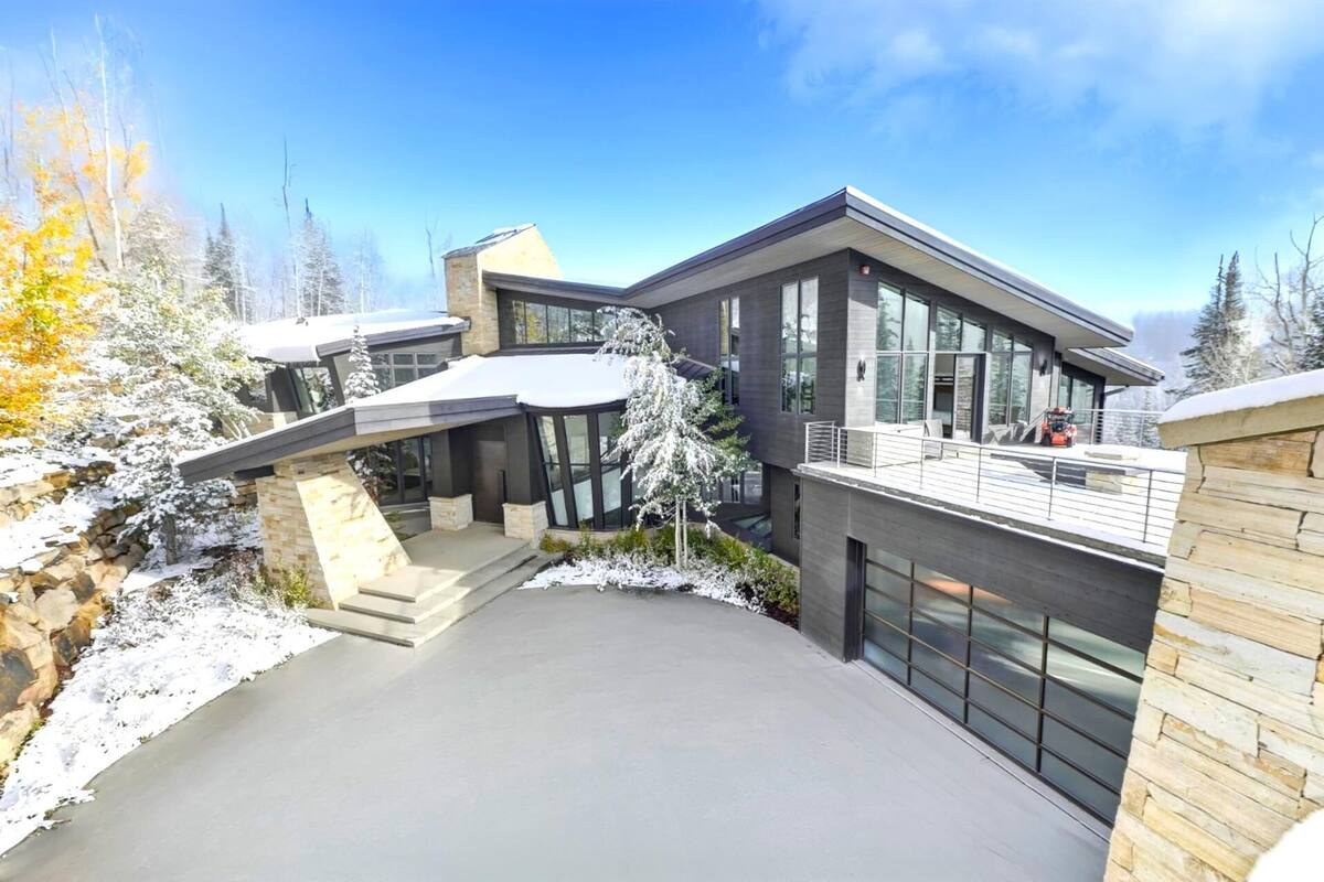 A modern multi-level house with a covered driveway, large windows, and a view of snow-covered trees.