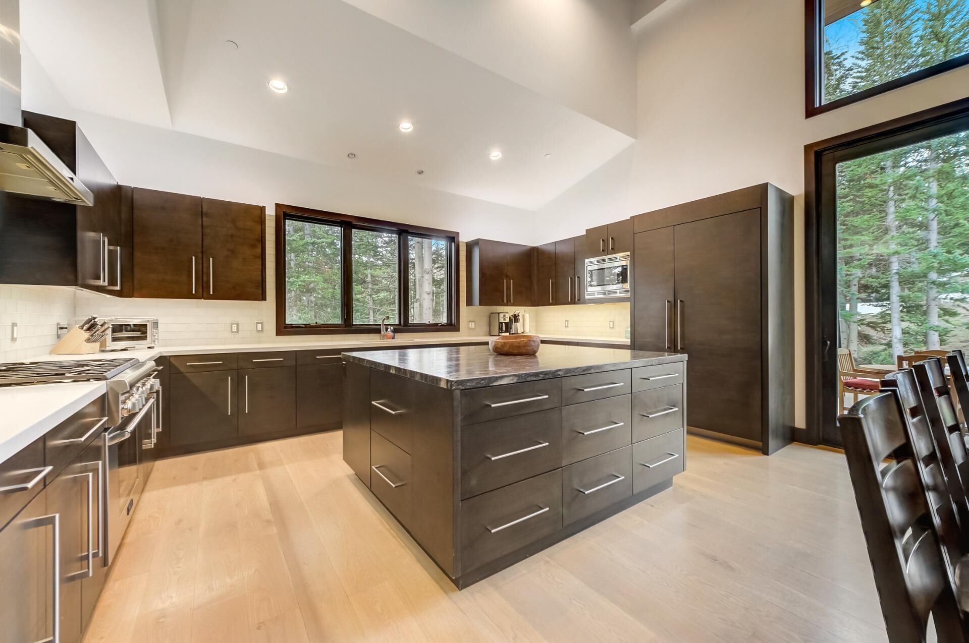 Une cuisine moderne avec des meubles de couleur sombre, un grand îlot central et une vue sur la forêt par les fenêtres.