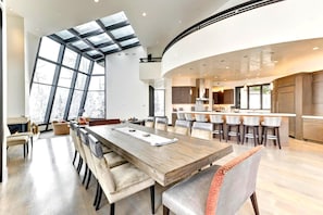 Un comedor amplio con una gran mesa de madera y múltiples sillas, una cocina con electrodomésticos modernos, y un ventanal que proporciona luz natural.