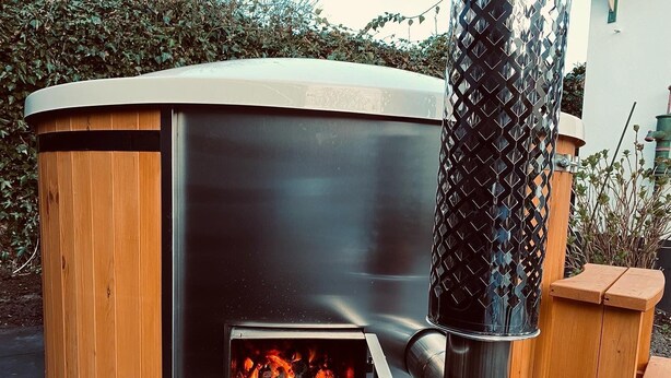 Un horno a leña con chimenea metálica, rodeado de construcciones de madera y follaje.