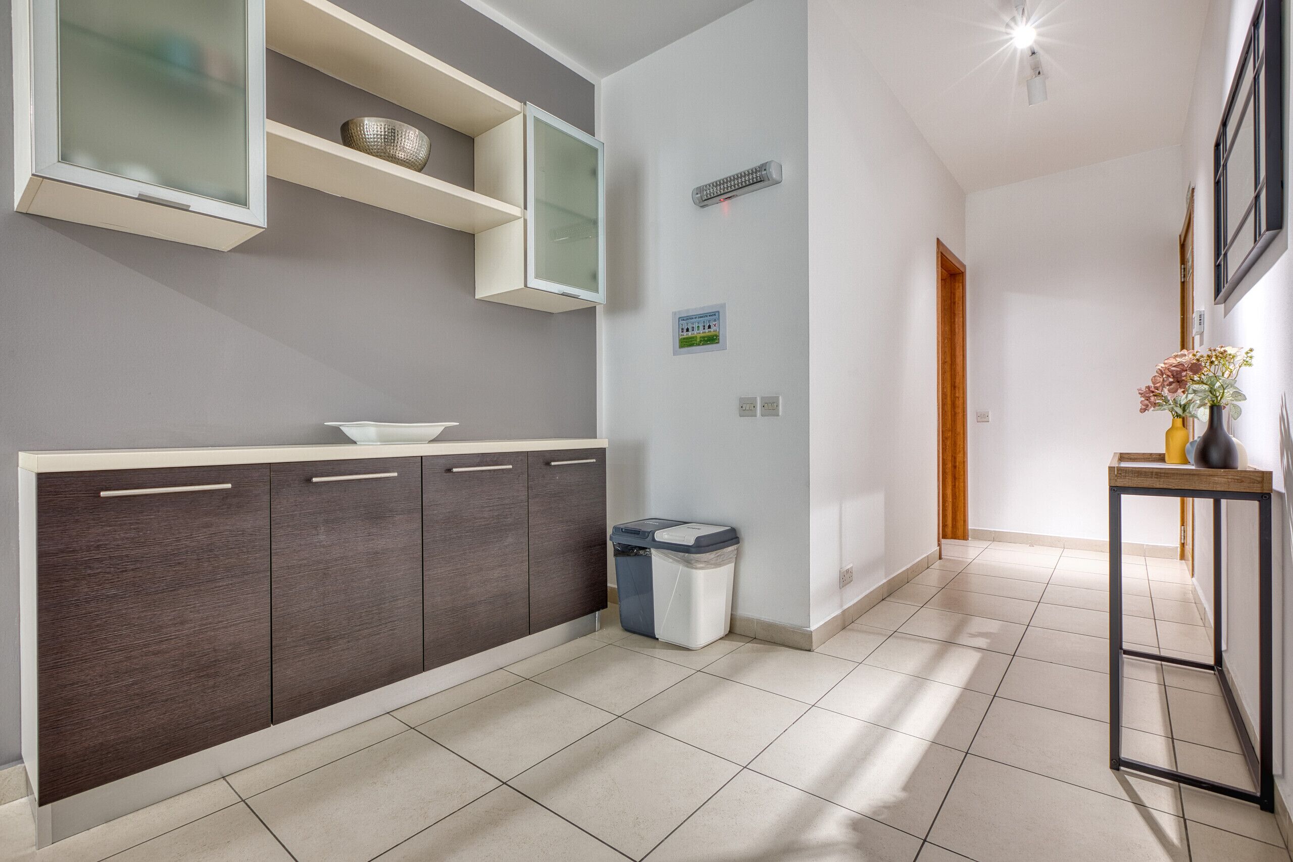 A modern kitchen with dark wood cabinets, a white countertop, and a built-in microwave. There is a trash bin and a small table with a vase of flowers.