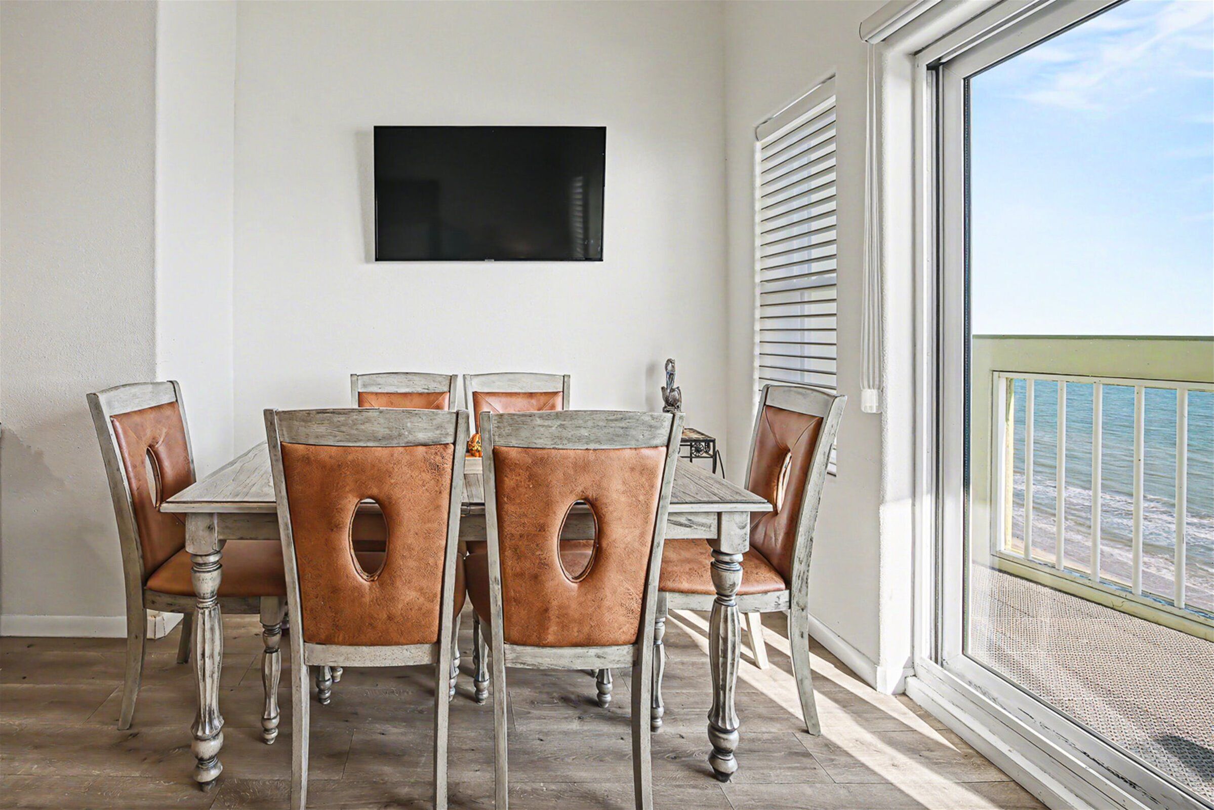 A dining area with a table and six chairs, a mounted flat-screen TV, and a view of the ocean through a balcony door.