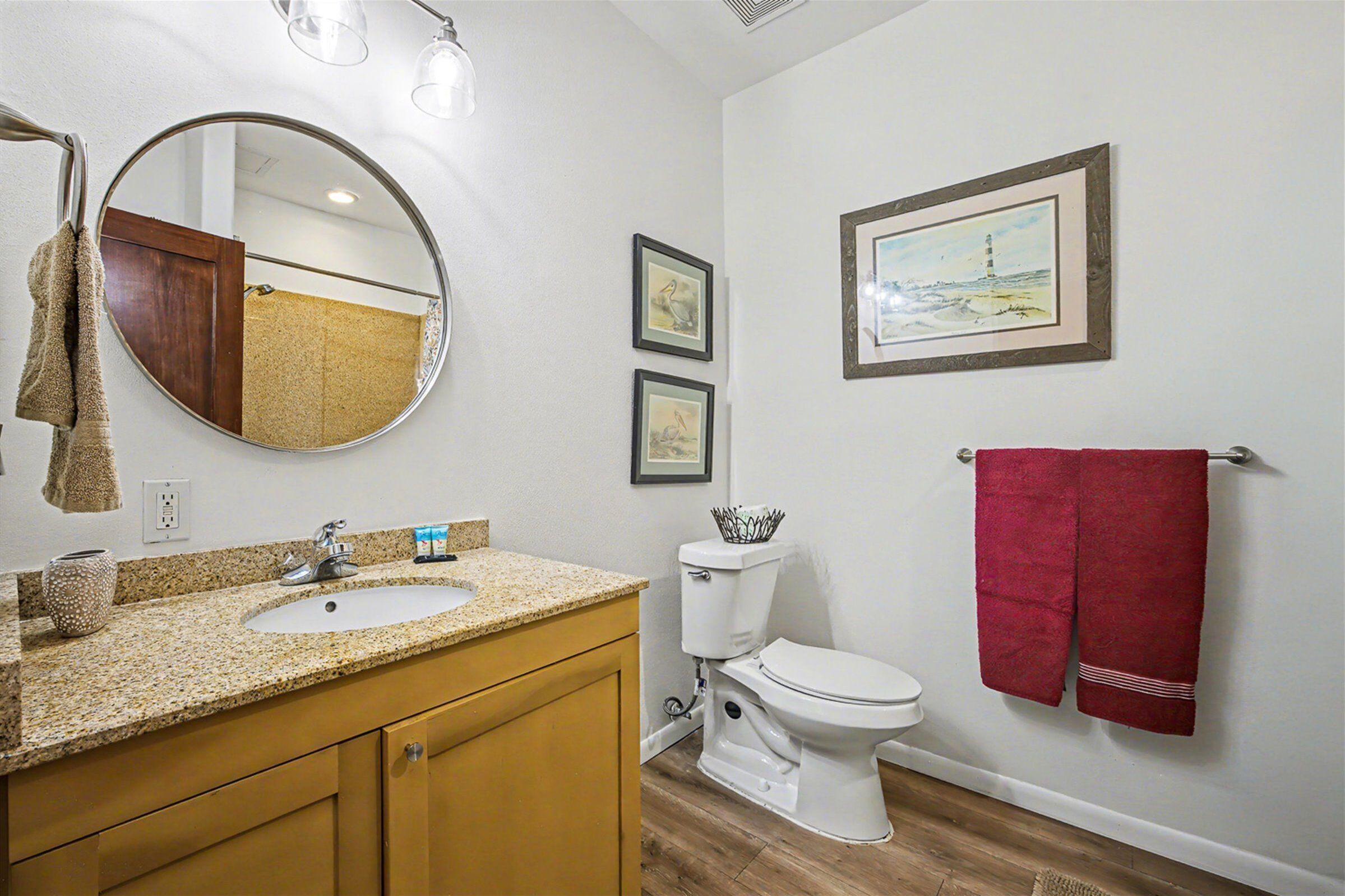 A bathroom with a granite countertop sink, wooden cabinets, a white toilet, and a red towel hanging on a towel rack. There are framed pictures on the wall and a mirror above the sink.