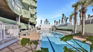 Un espace piscine avec une eau d’un bleu limpide, entouré de palmiers et un bâtiment à plusieurs niveaux doté de balcons.